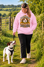 A woman walking a dog on a grassy path outdoors, wearing a light pink hoodie with a large graphic print that reads 'Dog Sister Chief Best Friend' and 'Peanuts in Garden'.
