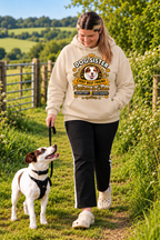 A woman walking a small white and brown dog on a grassy outdoor path. She is wearing a cream-colored hoodie with a 'Dog Sister' graphic print, black pants, and white clogs.