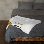 A white fleece blanket with a black and gold cartoon bird design is folded on a bed with gray bedding and pillows.